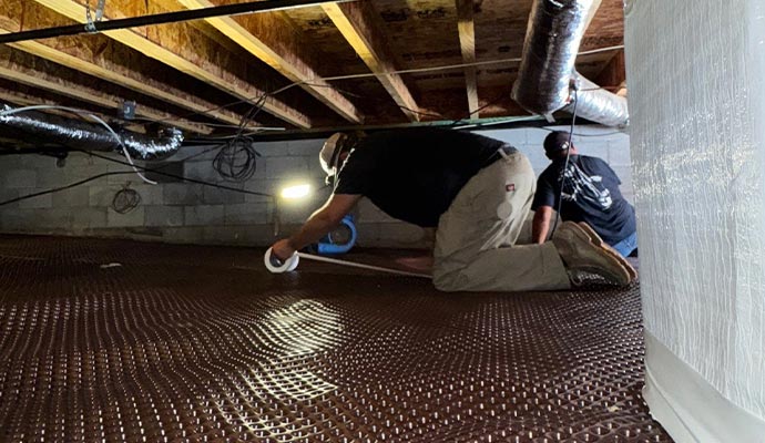 Crawlspace waterproofing and drainage mat installation Two workers installing a dimpled drainage mat in a crawlspace