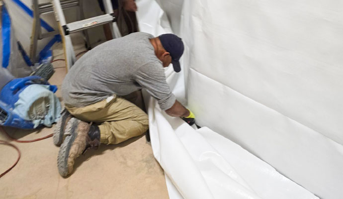 Technician installing a white waterproof vapor barrier liner along a basement wall