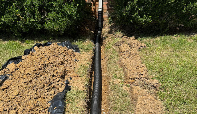 A trench in the yard with a black drainage pipe running through it