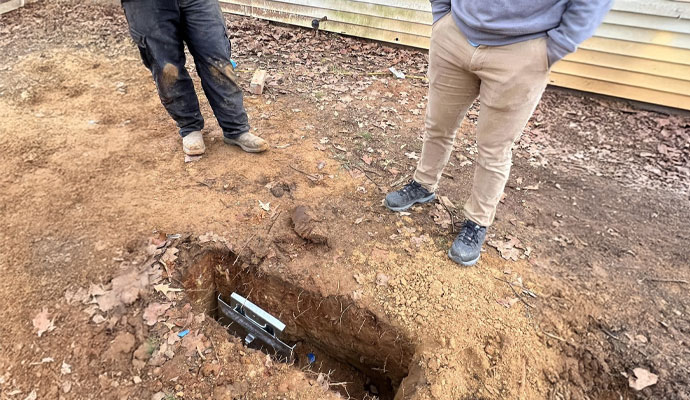Excavation for metal bracket installation for foundation stabilization Excavated hole with metal foundation bracket embedded in soil near building exterior