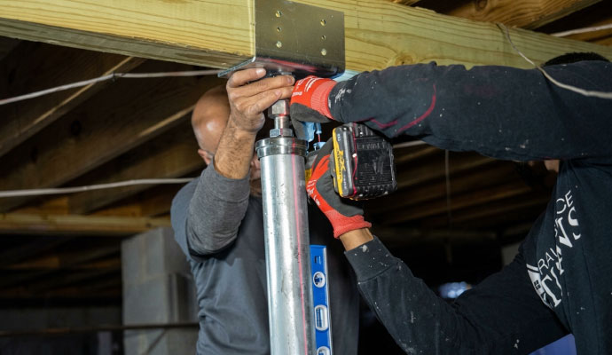 Technicians installing an adjustable steel power post beneath crawlspace floor joists